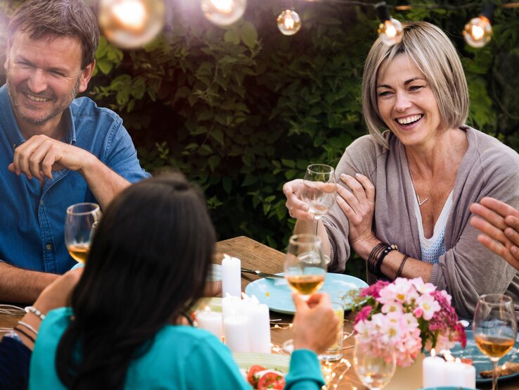 Eine Frau sitzt an einer Tafel bei einer Gartenparty und unterhält sich mit der Frau gegenüber. Die Runde sieht glücklich und entspannt aus.