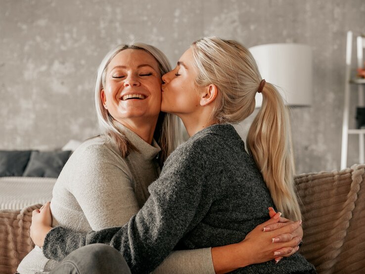 Eine ältere und eine jüngere Frau sitzen auf einer Couch und umarmen sich. Sie jüngere Frau gibt der älteren einen Kuss auf die Wange. | © Adobe Stock/ Vita