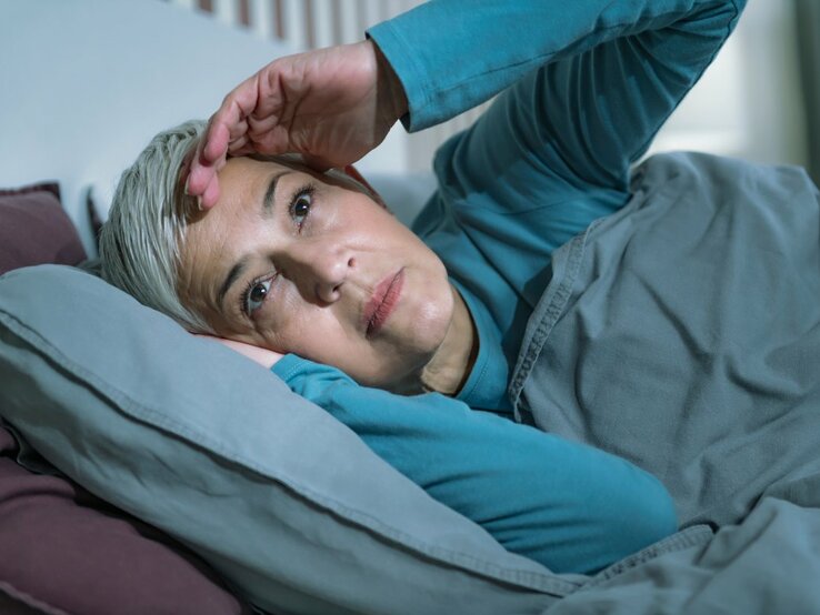 Das Bild zeigt eine ältere Frau mit kurzen grauen Haaren, die im Bett liegt und offenbar Schwierigkeiten beim Schlafen hat. Sie stützt ihren Kopf mit einer Hand, während sie nachdenklich oder besorgt nach oben schaut. Die Frau trägt ein türkisfarbenes Pyjamaoberteil und liegt unter einer grauen Bettdecke, die gut zu den lila Kissen passt. Das Bild könnte die Problematik der Schlafschwierigkeiten bei älteren Menschen illustrieren, die manchmal als senile Bettflucht bezeichnet werden.