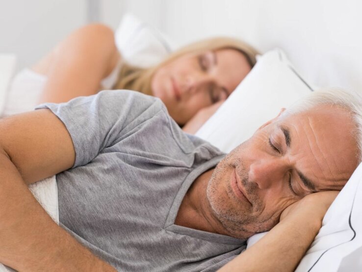 Das Bild zeigt ein älteres Paar, das friedlich im Bett schläft. Der Mann liegt im Vordergrund und scheint tief zu schlafen, mit einer Hand unter seinem Kopf und geschlossenen Augen. Er hat kurzes graues Haar und trägt ein graues T-Shirt. Die Frau liegt im Hintergrund und ist ebenfalls schlafend dargestellt, mit dem Kopf auf einem Kissen und dem Gesicht teils vom Bettrahmen verdeckt. Sie hat blondes Haar und trägt ein weißes Oberteil. Die Szene wirkt ruhig und entspannt, mit weicher Beleuchtung, die eine gemütliche Atmosphäre schafft. Es ist ein intimes und privates Bild, das Ruhe und Komfort in einem häuslichen Umfeld darstellt.