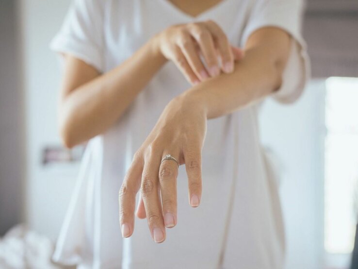  Das Bild zeigt eine Person, die ihren eigenen Unterarm betrachtet und mit der anderen Hand leicht darüber streicht oder kratzt. Das Individuum trägt ein weißes T-Shirt und hat einen Ring am Finger, was darauf hindeuten könnte, dass es sich um eine Frau handelt. Die Aufmerksamkeit auf den Arm und die Geste des Betrachtens oder Kratzens könnten auf Hautprobleme wie Nesselsucht hinweisen, bei der die Betroffenen oft einen Juckreiz oder eine Reizung verspüren. 