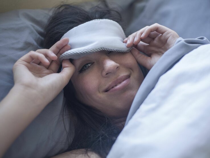 Eine Frau liegt im Bett. Eine Schlafmaske verdeckt ein Auge. Sie lächelt freundlich den Betrachter des Bildes an.