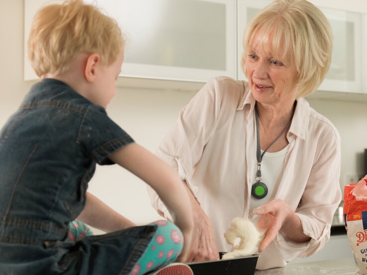 Eine ältere Frau mit blondem Haar knetet Teig und lächelt ein blondes Kind an, das in bunter Leggings auf der Küchenarbeitsplatte sitzt.