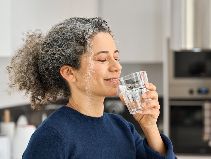 Titelbild Staatl. Fachingen Eine Frau mit grauen Locken und blauem Pullover trinkt lächelnd ein Glas Wasser in einer modernen Küche.