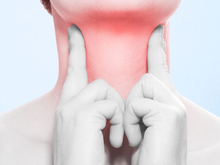 Nahaufnahme eines Halses vor hellem, bläulich wirkendem Hintergrund; beide Hände in Graustufen legen die Zeigefinger links und rechts an den Kehlbereich. Der Hals ist rötlich eingefärbt und betont den Bereich der Mandeln, um Schmerzen, Reizung oder Beschwerden durch Mandelsteine zu veranschaulichen. Der Bildausschnitt zeigt keine Gesichtszüge und konzentriert sich ausschließlich auf den Rachenbereich als medizinische Illustration.