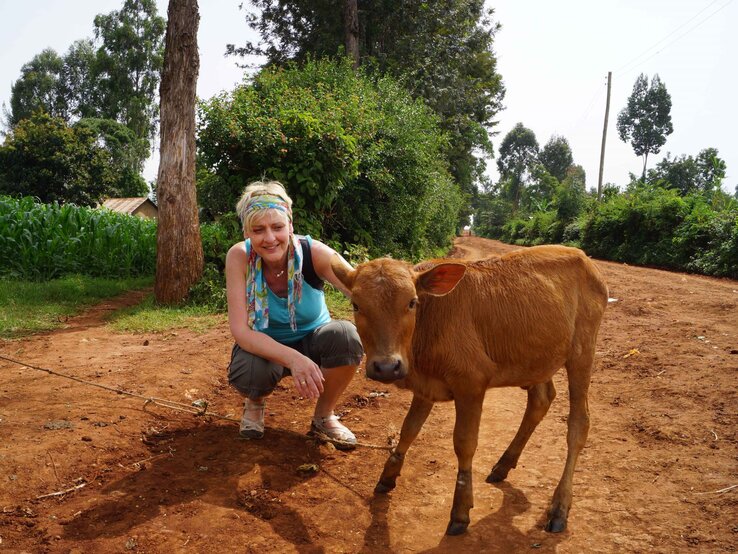 Preisträgerin Christina Schreiber mit Kuh | © GOLDENE BILD der FRAU Eine lächelnde Frau mit bunter Haarbinde hockt neben einem braunen Kalb auf einem roten Sandweg vor dichtem Grün. | © GOLDENE BILD der FRAU