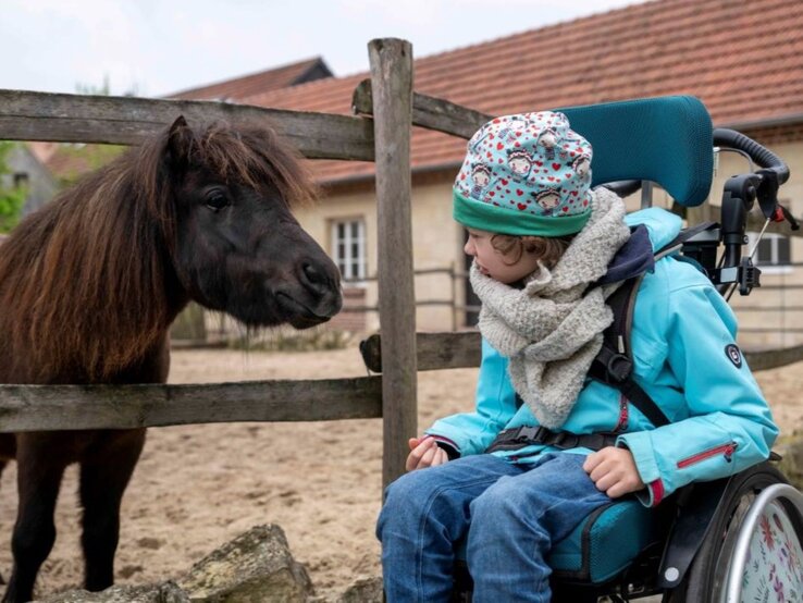 Hof Feismann | © GOLDENE BILD der FRAU Kind im Rollstuhl mit bunter Mütze und Winterjacke blickt neugierig ein schwarzes Pony an, das hinter einem Holzzaun steht. | © GOLDENE BILD der FRAU