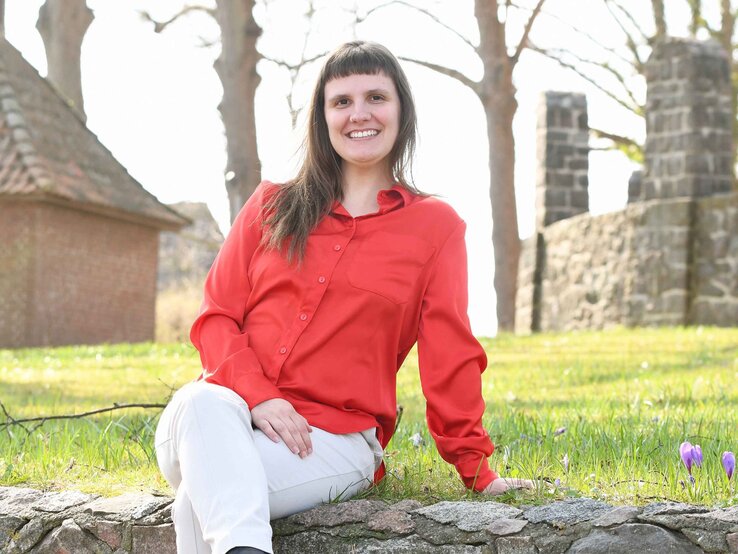 Preisträgerin Miriam Peters Junge Frau mit braunen Haaren, roter Bluse und weißer Hose sitzt lächelnd auf einer Mauer vor alten Steinruinen im Sonnenschein.