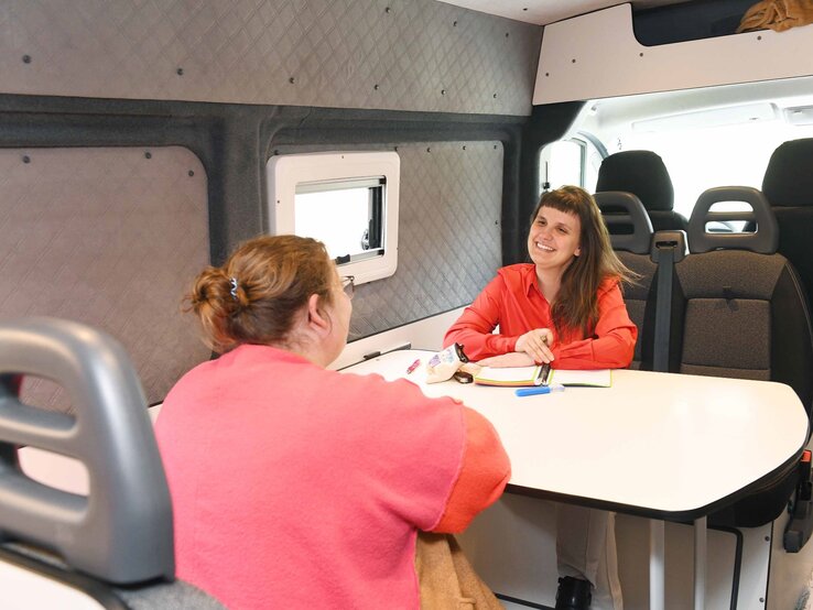 Miriam Peters bei der Beratung | © GOLDENE BILD der FRAU Zwei Frauen unterhalten sich freundlich an einem Tisch in einem Camper, eine schreibt lächelnd in ein Notizbuch. | © GOLDENE BILD der FRAU