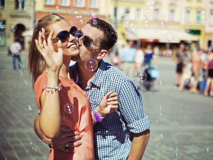 Ein Paar ist auf der Straße umgeben von Seifenblasen. Er küsst sie auf die Wange. Sie lächelt.