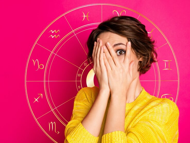 sternzeichen-schlechter-ruf Eine überraschte Frau in einem gelben Oberteil hält ihre Hände vor ihr Gesicht und späht durch ihre Finger. Im Hintergrund ist eine leuchtend rosa Grafik mit astrologischen Symbolen, darunter das Rad des Tierkreises mit den Zeichen von Widder bis Fische.