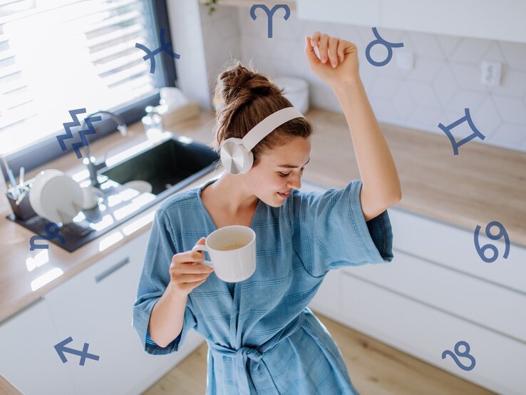 Eine junge Frau in einem entspannten blauen Kleid mit weißen Kopfhörern tanzt in einer hellen Küche und hält eine Tasse Kaffee. Sie scheint glücklich und energiegeladen zu sein. Über das Bild verteilt sind verschiedene Astrologie-Symbole wie Zwillinge, Wassermann, Steinbock und Krebs, die darauf hindeuten könnten, dass das Bild sich mit Astrologie oder Horoskopen beschäftigt.