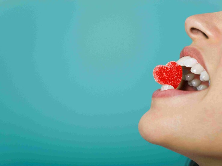 Frau hält herzförmige Süßigkeit zwischen den Zähnen. | © PhotoAlto/Laurence Mouton/Getty Images