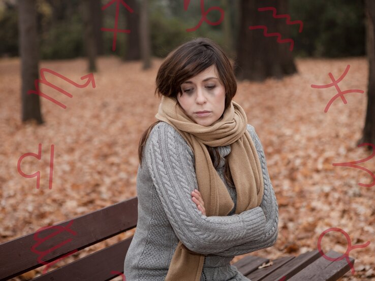 sternzeichen-herbstdepression Eine nachdenkliche Frau sitzt mit verschränkten Armen auf einer Parkbank an einem Herbsttag. Sie trägt einen grauen Strickpullover und einen beigen Schal. Um sie herum liegen gefallene Blätter. Im Hintergrund sind Symbole von Sternzeichen zu sehen, die in Rot über das Bild gelegt sind. Die Frau wirkt bedrückt oder nachdenklich, möglicherweise in Verbindung mit einer Herbstdepression.