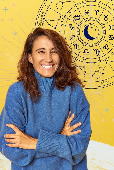 Eine lächelnde Frau mit langen brünetten Haaren steht vor einem gelben Hintergrund und trägt einen blauen Pullover. Sie ist gut gelaunt. Hinter ihr ist der Tierkreis zu sehen mit den 12 astrologischen Zeichen. Daneben sind grafische Strahlen und zarte bunte Sterne zu sehen. Die Grundstimmung ist positiv und optimistisch.