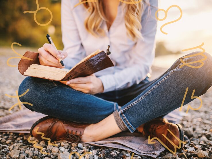 Eine Frau sitzt im Schneidersitz an einem Steinstrand und macht sich Notizen in einem Buch.