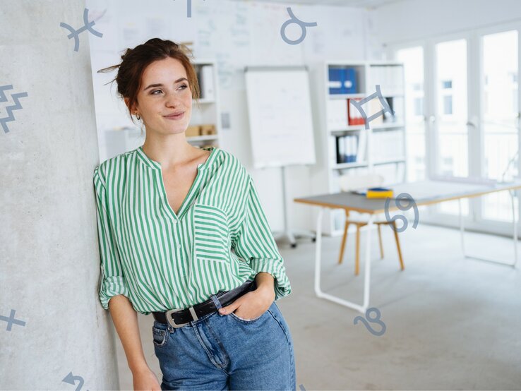 Eine Frau mit weiß-grüner Bluse steht lächelnd an einer Wand. Hinter ihr sieht man ihren Arbeitsplatz.
