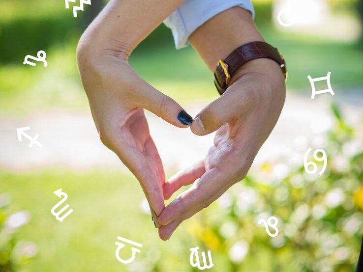Die Hand einer Frau und eines Mannes formen ein Herz. Im Hintergrund ist das Grün von Natur zu sehen. Umgeben sind die Hände von den astrologischen Zeichen des Tierkreises. 