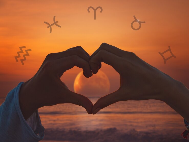 Zwei Hände formen bei Sonnenuntergang ein Herz um eine aufgehende Vollmond-Silhouette am Meer. Im Hintergrund sind astrologische Symbole, einschließlich des Symbols für den Wassermann, Stier, Schütze und Zwillinge, über einen orangefarbenen Himmel verteilt.