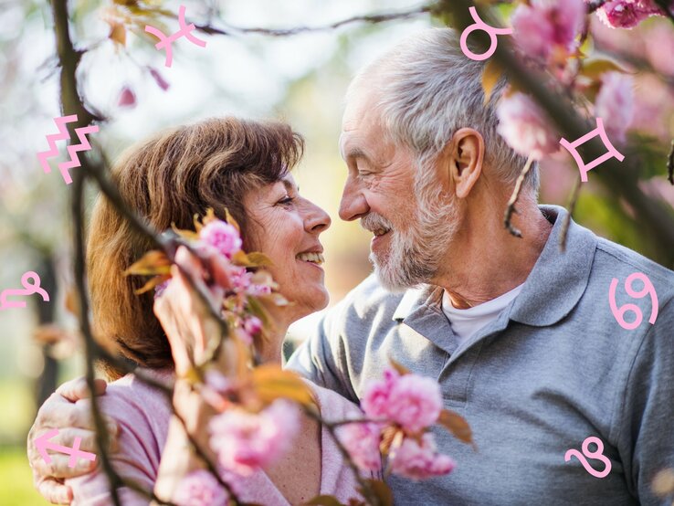 Ein älteres Paar, das liebevoll und lächelnd Augenkontakt hält. Die Frau hat braunes Haar und trägt ein rosa Oberteil, während der Mann graues Haar und einen grauen Pullover hat. Sie umarmen sich inmitten der Blüten eines Kirschblütenbaums. Grafische Symbole der Tierkreiszeichen sind über das Bild verteilt, was auf einen astrologischen oder romantischen Kontext hindeutet.