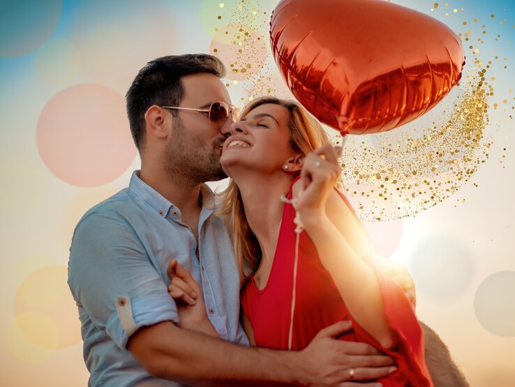 Auf dem Bild ist ein glückliches Paar zu sehen, das sich liebevoll umarmt und küsst. Der Mann trägt ein hellblaues Hemd und eine Sonnenbrille, während die Frau ein leuchtend rotes Kleid trägt und ebenfalls eine Sonnenbrille hat. Sie hält einen herzförmigen, roten Luftballon. Im Hintergrund sind verschwommene Lichtkreise und eine Glitzerwolke, die den feierlichen und romantischen Anlass unterstreicht. Das Bild strahlt Wärme und Zuneigung aus, verstärkt durch das weiche, warme Licht der Umgebung.