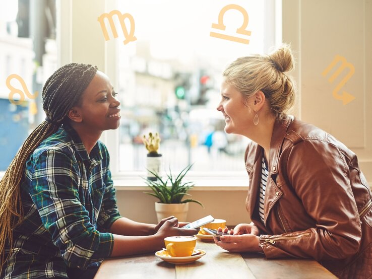 Zwei Frauen sitzen an einem Tisch in einem Café und unterhalten sich fröhlich. Die Frau links trägt ein kariertes Hemd und hat geflochtene Zöpfe, die Frau rechts trägt eine braune Lederjacke und hat blonde Haare zu einem Knoten gebunden. Im Hintergrund sind draußen unscharf Gebäude und Menschen zu sehen. Um sie herum sind astrologische Symbole von verschiedenen Sternzeichen platziert.