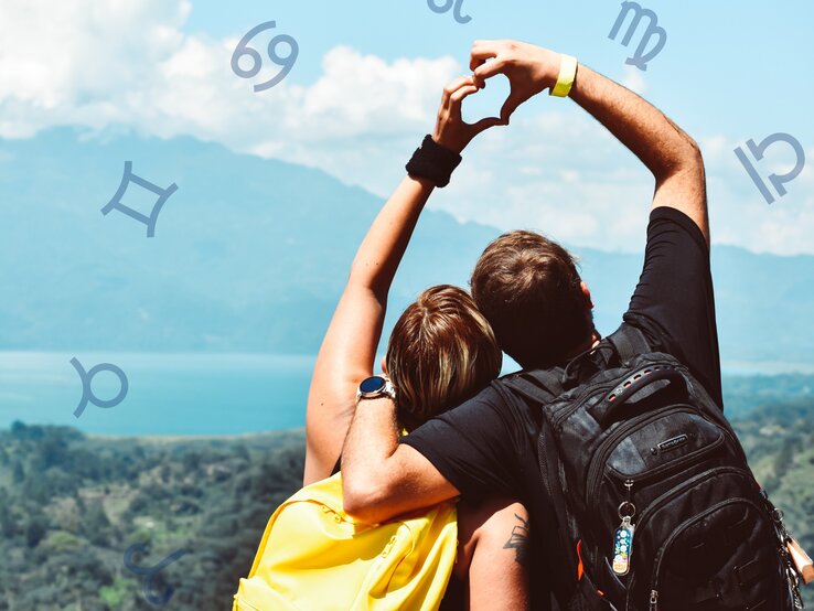 Ein Paar steht mit dem Rücken zur Kamera und blickt auf eine wunderschöne Landschaft mit Bergen und einem See. Beide tragen Rucksäcke und formen mit ihren Armen ein Herz. Um sie herum sind astrologische Symbole von verschiedenen Sternzeichen platziert.