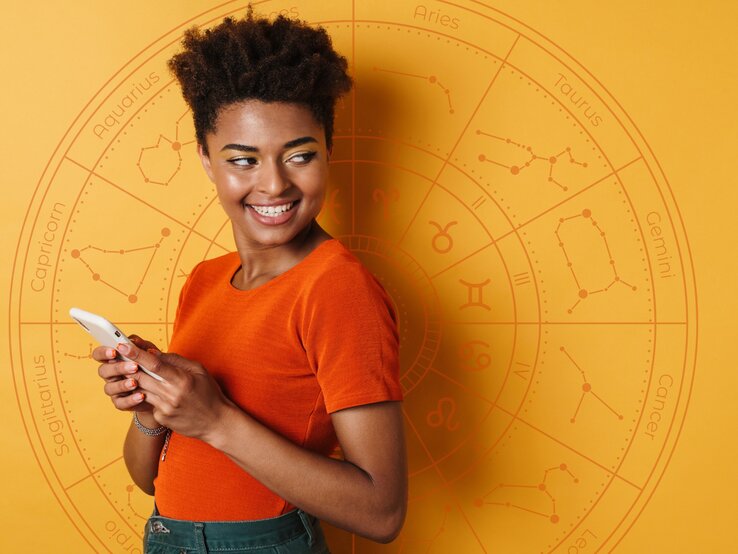 Eine Frau in einem orangefarbenen Oberteil hält ein Smartphone und lächelt. Sie steht vor einem orangefarbenen Hintergrund mit einem astrologischen Rad und Sternzeichen-Symbolen.
