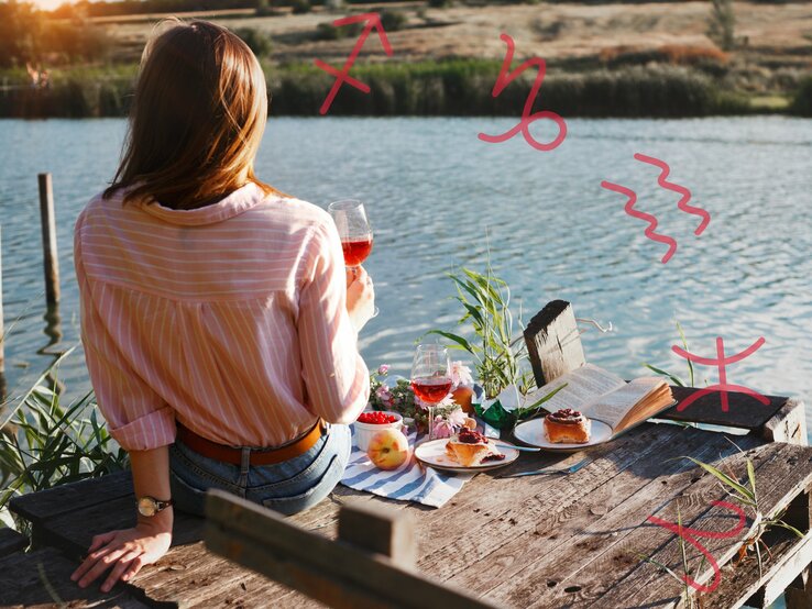 Eine Frau sitzt mit dem Rücken zur Kamera auf einem hölzernen Steg am Wasser. Sie trägt ein rosafarbenes, gestreiftes Hemd und hält ein Glas Roséwein in der Hand. Vor ihr auf dem Steg liegen ein aufgeschlagenes Buch, zwei Teller mit Kuchen, ein weiteres Glas Wein, eine Schale mit Beeren und ein Pfirsich. Im Hintergrund ist eine ruhige, ländliche Landschaft zu sehen. In der Luft sind verschiedene astrologische Symbole gezeichnet: Schütze, Steinbock, Wassermann, Fische und Widder.