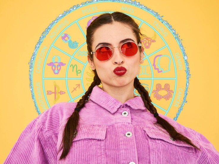 Eine Frau mit geflochtenen Haaren trägt eine rot getönte Sonnenbrille und ein lila Cordhemd. Sie macht einen spielerischen 'Duckface'-Ausdruck. Hinter ihr befindet sich ein buntes Tierkreisrad mit Symbolen, die die zwölf Sternzeichen darstellen, vor einem gelben Hintergrund.