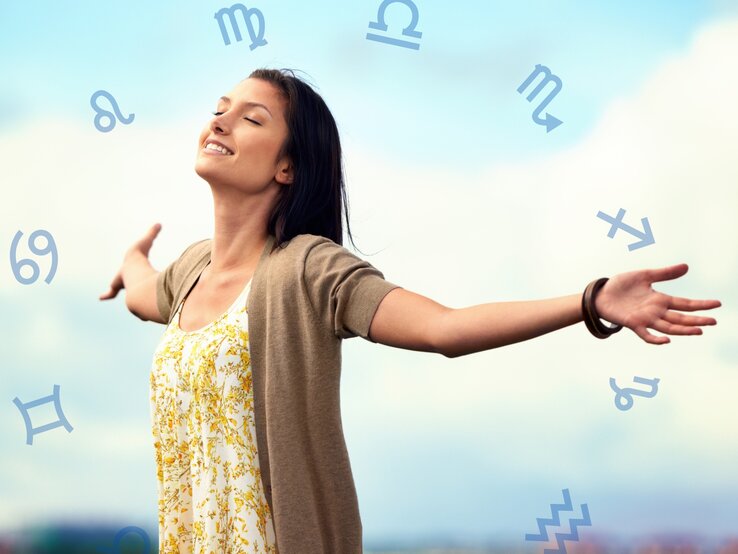 Eine lächelnde Frau steht mit ausgebreiteten Armen im Freien unter einem blauen Himmel. Sie trägt ein weißes Kleid mit gelbem Blumenmuster und eine beige Strickjacke. Um sie herum schweben verschiedene astrologische Symbole, die auf den Tierkreis hinweisen. Die Frau wirkt glücklich und frei.