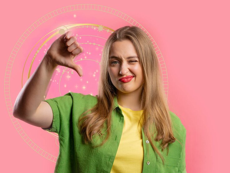 Eine junge Frau mit langen, blonden Haaren steht vor einem rosa Hintergrund mit einem astrologischen Rad. Sie trägt ein grünes Hemd über einem gelben T-Shirt und schaut mit einem unzufriedenen Gesichtsausdruck in die Kamera. Ihre Lippen sind rot geschminkt, und sie zeigt mit ihrem Daumen nach unten, als Zeichen der Missbilligung.