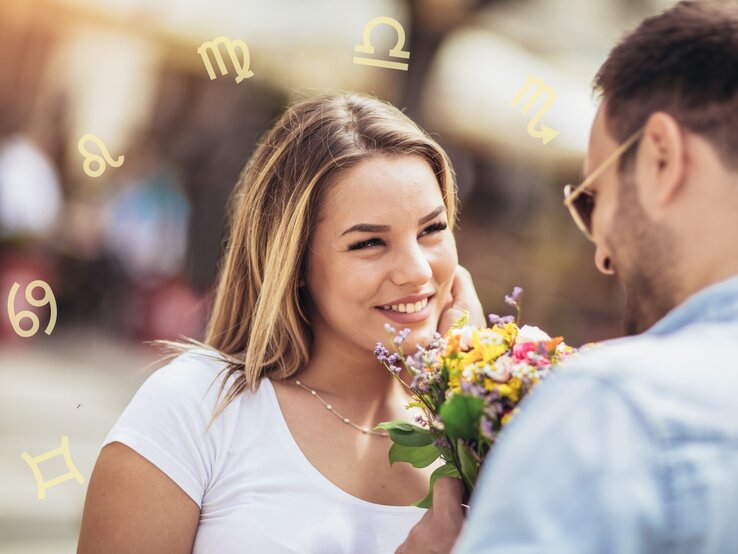 Ein Mann und eine Frau stehen sich gegenüber und lächeln sich an. Der Mann hält einen Blumenstrauß in der Hand, den er der Frau überreicht. Im Hintergrund sind verschwommene Menschen und Gebäude zu sehen. Um die Köpfe der beiden Personen sind verschiedene astrologische Symbole angeordnet, die auf die Sternzeichen hinweisen. Die Frau trägt ein weißes T-Shirt und hat blonde Haare, während der Mann eine Sonnenbrille und ein blaues Hemd trägt.
