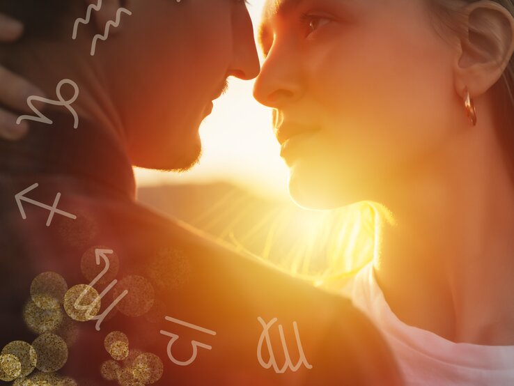 Ein Mann und eine Frau stehen sich sehr nah gegenüber, beinahe in einer Umarmung, und schauen sich intensiv in die Augen. Die Szene wird von warmem, goldenem Sonnenlicht durchflutet, das von hinten auf die beiden Personen fällt und eine romantische Atmosphäre schafft. Um sie herum sind astrologische Symbole verteilt, die auf verschiedene Sternzeichen hinweisen. Der Mann hat einen leichten Bart und trägt eine dunkle Jacke, während die Frau goldene Creolen trägt und ein weißes Oberteil.