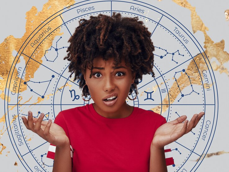 Eine junge Frau mit dunklen lockigen Haaren und einem roten T-Shirt hebt verwirrt beide Hände und blickt mit einem erstaunten Gesichtsausdruck in die Kamera. Im Hintergrund befindet sich ein astrologischer Kreis mit den Symbolen und Konstellationen der zwölf Tierkreiszeichen in Blau auf einem grauen Hintergrund, verziert mit goldenen Akzenten.