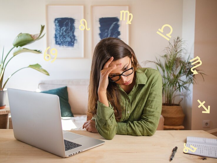 Das Bild zeigt eine Frau, die vor einem Laptop an einem Schreibtisch sitzt und erschöpft oder gestresst wirkt. Sie stützt ihren Kopf mit einer Hand und trägt eine Brille sowie ein grünes Hemd. Auf dem Tisch liegen ein Notizbuch und ein Stift. Im Hintergrund sind Zimmerpflanzen und zwei gerahmte Bilder an der Wand zu sehen. Um sie herum schweben verschiedene astrologische Symbole, was auf einen Zusammenhang mit Astrologie oder Sternzeichen hinweist. Die Szene vermittelt eine Atmosphäre von Müdigkeit oder Überforderung in einer häuslichen oder Arbeitsumgebung.