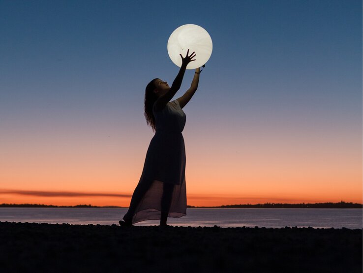 Das Bild zeigt eine Frau im langen Kleid, die am Strand steht und ihre Arme hochhält, als würde sie den großen, leuchtenden Vollmond über ihrem Kopf halten. Der Himmel im Hintergrund ist während des Sonnenuntergangs in wunderschönen Blau- und Orangetönen gefärbt, was eine ruhige und magische Atmosphäre schafft. Die Silhouette der Frau und die Reflexion des Lichts auf dem Wasser verstärken die mystische Stimmung des Bildes. Es vermittelt den Eindruck von Harmonie und Verbindung mit der Natur, passend zu einem Mondhoroskop-Thema.