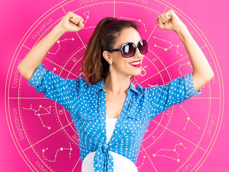 Eine Frau in einer blauen, gepunkteten Bluse posiert selbstbewusst vor einem astrologischen Rad, das verschiedene Sternzeichen zeigt. Sie trägt eine Sonnenbrille und hebt ihre Arme in einer kraftvollen Pose. Das Bild vermittelt Optimismus, Selbstbewusstsein und Erfolg.