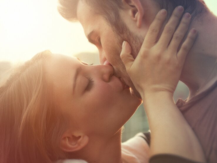 Ein Paar, bestehend aus einer Frau und einem Mann, teilt einen innigen Kuss. Die Frau hält das Gesicht des Mannes sanft mit beiden Händen, während die Sonne im Hintergrund für eine warme, romantische Beleuchtung sorgt.