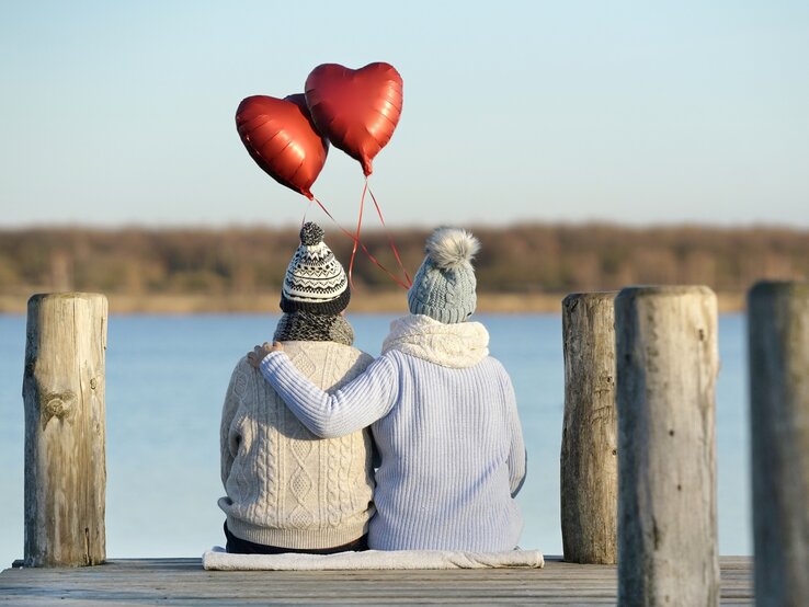 Zwei Personen sitzen eng umschlungen auf einem Holzsteg an einem See, mit Blick auf das Wasser. Sie tragen warme Winterkleidung und halten drei rote Herzballons in der Hand, die in die Luft steigen. Die Szene vermittelt eine ruhige und romantische Atmosphäre.