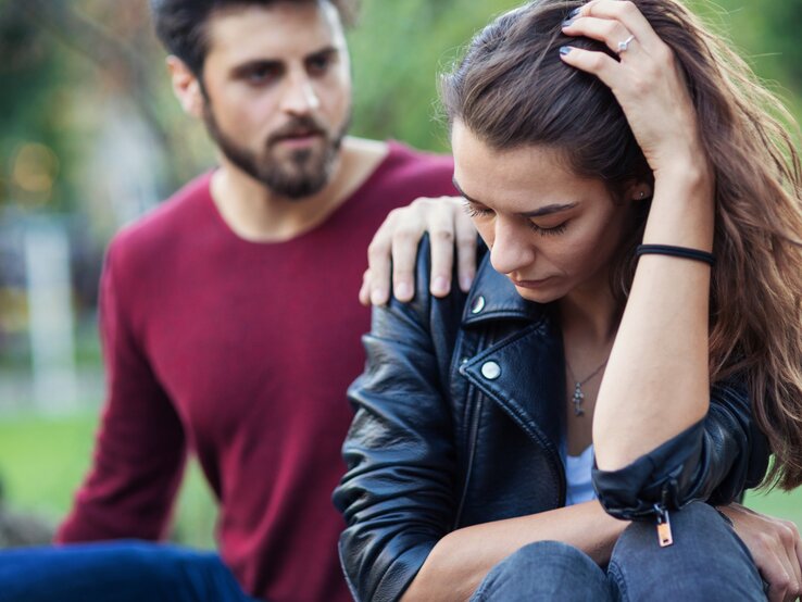 kw-44.jpg Ein Mann legt tröstend seine Hand auf die Schulter einer nachdenklich und traurig wirkenden Frau. Beide sitzen im Freien, die Frau hält ihren Kopf mit einer Hand und wirkt besorgt, während der Mann mitfühlend neben ihr sitzt.