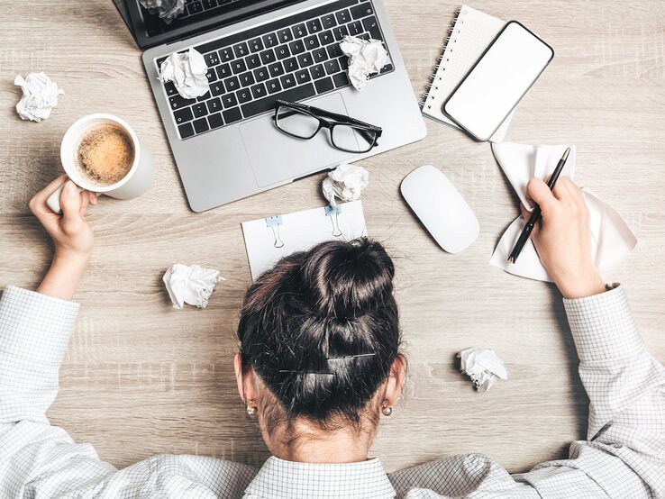 Eine Person liegt erschöpft mit dem Kopf auf einem Stapel von Formularen und Dokumenten, darüber ist die Tastatur eines Laptops zu sehen. Die Szene vermittelt Stress oder Überforderung durch Arbeit oder Bürokratie.