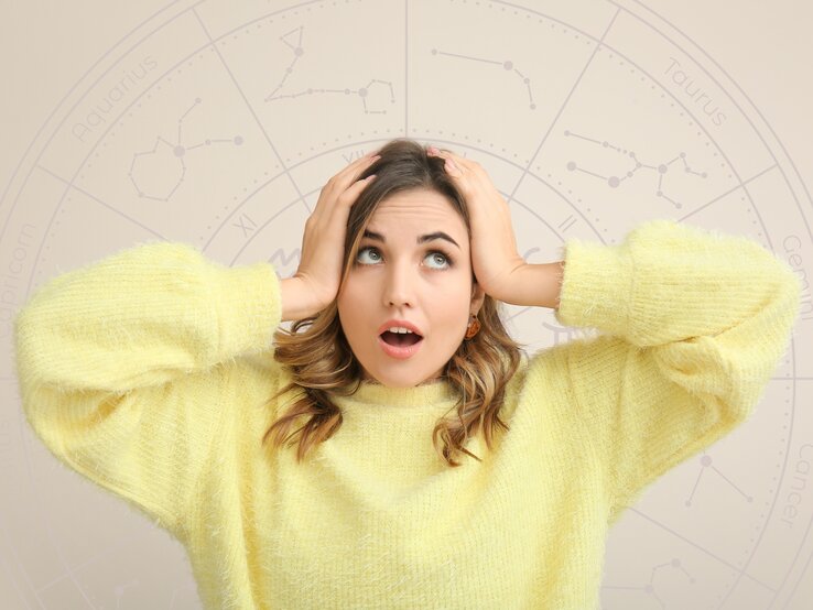 Frau in gelbem Pullover schaut überrascht und hält sich die Hände an den Kopf. Im Hintergrund ist ein astrologisches Rad mit Tierkreiszeichen wie Aquarius, Taurus und Gemini zu sehen.