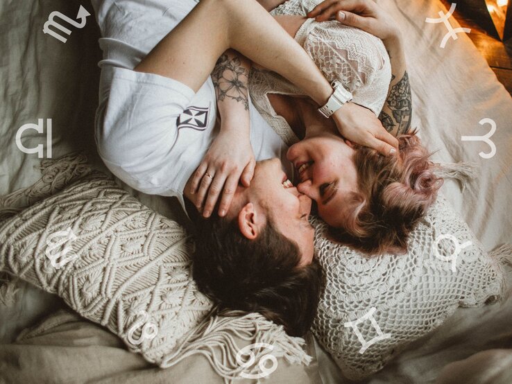 Ein verliebtes Paar liegt eng umschlungen auf einem Bett mit gemütlichen, gehäkelten Kissen. Beide lächeln sich an, während um sie herum astrologische Symbole der Tierkreiszeichen schweben. Die Szene strahlt Intimität, Romantik und eine harmonische Atmosphäre aus.