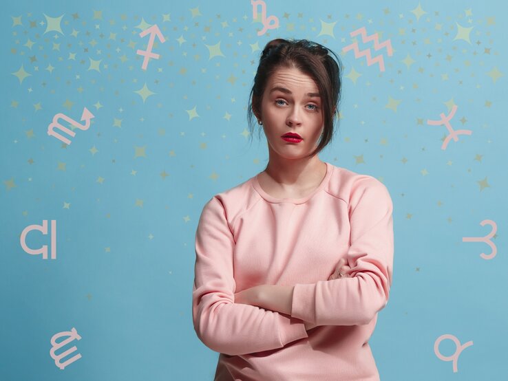 Eine Frau in einem rosa Pullover steht vor einem hellblauen Hintergrund mit pastellfarbenen Sternzeichen-Symbolen und goldenen Sternen. Sie hat die Arme verschränkt und einen nachdenklichen Gesichtsausdruck, der Fragen oder Unsicherheiten ausdrücken könnte. Das Bild symbolisiert astrologische Themen und persönliche Reflektion im Zusammenhang mit den Sternzeichen.