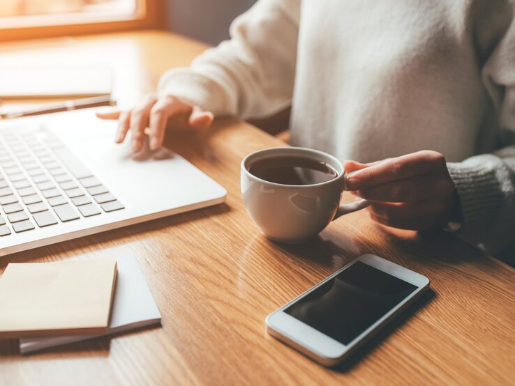Eine Person sitzt an einem Holztisch und arbeitet an einem Laptop. In der rechten Hand hält sie eine weiße Tasse mit heißem Kaffee, während die linke Hand die Tastatur bedient. Neben dem Laptop liegen Notizzettel und ein Stift. Auf dem Tisch befindet sich außerdem ein Smartphone mit schwarzem Bildschirm. Durch ein Fenster im Hintergrund fällt warmes, natürliches Licht und erzeugt eine gemütliche Atmosphäre.