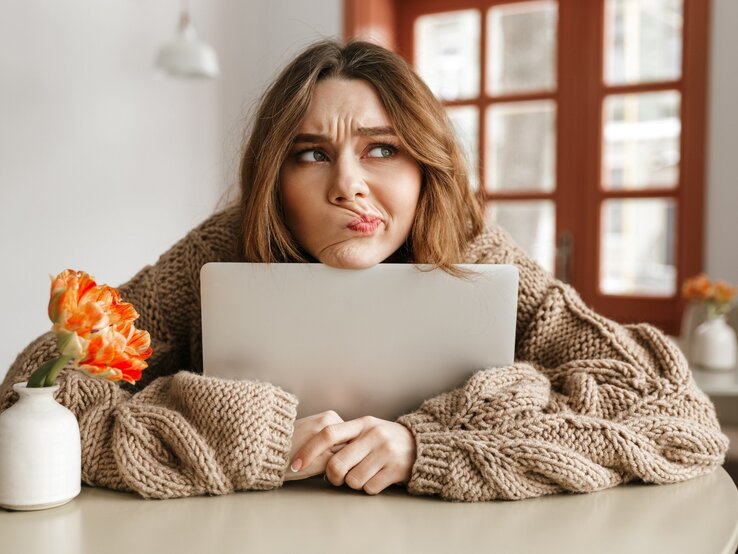 Eine nachdenkliche Frau stützt ihr Kinn auf einen Laptop, während sie einen skeptischen Gesichtsausdruck zeigt. Sie trägt einen dicken, beige gestrickten Pullover und sitzt an einem Tisch. Links von ihr steht eine weiße Vase mit einer orangefarbenen Blume. Im Hintergrund sind Fenster mit Holzrahmen zu sehen, durch die Tageslicht hereinfällt. Die Szene vermittelt eine gemütliche, aber nachdenkliche Stimmung.