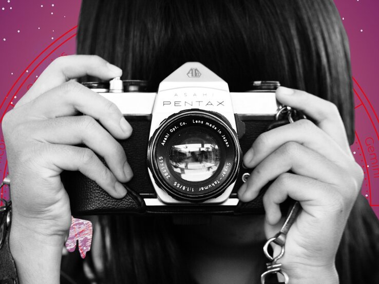 Eine Person hält eine analoge Kamera vom Typ Asahi Pentax vor das Gesicht, bereit zum Fotografieren. Im Hintergrund ist ein stilisiertes astrologisches Rad mit Sternzeichen auf violettem Grund zu sehen – Symbol für den Blick in die Zukunft und das Zusammenspiel von Kreativität und kosmischem Einfluss.