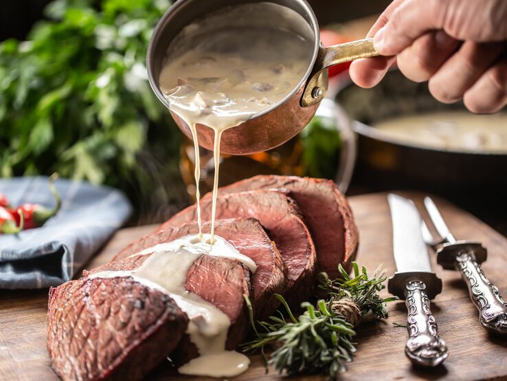 Eine Hand gießt cremige Sauce aus einem kleinen, antiken Kupfertopf über ein saftig aussehendes Stück rosa gebratenes Rindfleisch, das auf einem Holzbrett angerichtet ist. Neben dem Fleisch liegen frische Rosmarinzweige und silberne Besteckteile mit verzierten Griffen. 