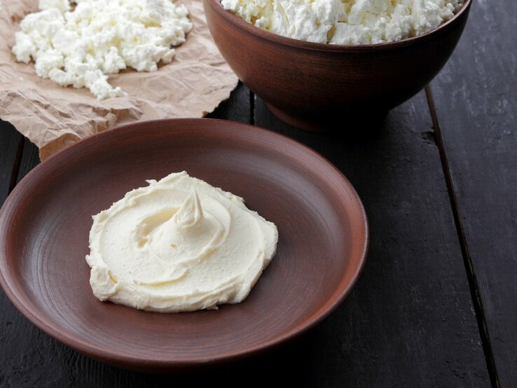 Ein Schlag frischer Quark oder Topfen ist kunstvoll auf einem dunkelbraunen Keramikteller serviert, der auf einer schwarzen Holzoberfläche steht. Im Hintergrund, außerhalb des Fokus, ist eine größere Menge desselben Produkts zu sehen, die in einer dunkelbraunen Keramikschüssel auf bewährtem Packpapier präsentiert wird. Die Farben und Texturen unterstreichen die Einfachheit und Natürlichkeit des Lebensmittels